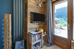 Een kamer met een wandtelevisie, houten lambrisering, een boekenplank vol boeken en uitzicht op een groen landschap door een deur.