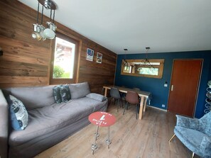 A room with a wooden wall, a grey sofa, a dining table with chairs, and a red stool. - Apartment 'Residence - Le Regain' with Mountain View, Private Terrace and Wi-Fi (Le Grand-Bornand)