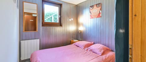 Un dormitorio con cama, ventana, radiador, mesita de noche y luz empotrada en la pared.