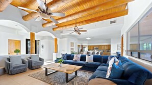 Un vaste espace de vie comprenant un grand canapé bleu, deux fauteuils, une table basse en bois et un ventilateur de plafond.