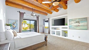 Une chambre à coucher avec un lit, un ventilateur de plafond, une télévision, des œuvres d’art et une vue sur des palmiers.