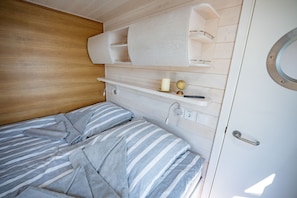 Ein Bett mit gestreifter Bettwäsche, ein Holz-Kopfteil, eine weiße Wand mit Einbauschränken und eine Tür mit rundem Fenster.