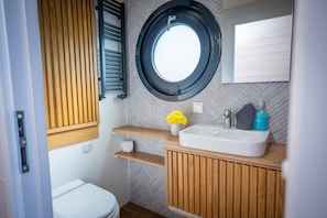 A compact bathroom with a toilet, a wooden vanity with a sink, a mirror, and a window. - House Boat 'Woma D12 Dawn' with Lake View, Wi-Fi and Air Conditioning (Fürstenberg/Havel)