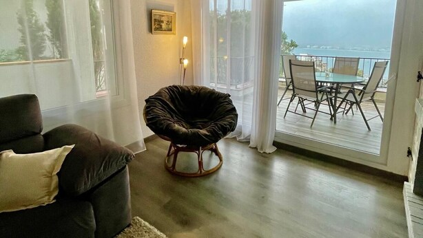 A living room with a dark sofa, a round chair, a small table, and a view of the sea through large windows. - Roses Apartment (Spain) with Sea and Mountain Views (Roses, Girona)