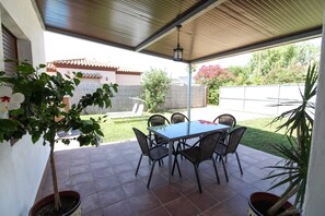 Um pátio externo com mesa e cadeiras, cercado por plantas em vasos e vista para uma área residencial.