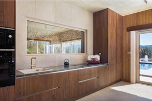 A modern kitchen with a stone backsplash, wooden cabinets, a built-in oven, and a large window overlooking a pool. - Villa Fantažija (Dicmo)