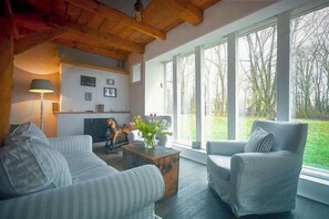 A living room with a sofa, armchair, and coffee table, a fireplace, and large windows with a view of trees. - Restored estate with sauna in Nordkehdingen (Oederquart)