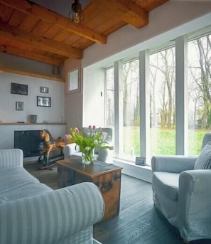 A living room with a fireplace, large windows, and a view of the outdoors. - Restored estate with sauna in Nordkehdingen (Oederquart)
