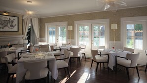 Comedor con varias mesas y sillas, ventanas amplias con cortinas y un candelabro.