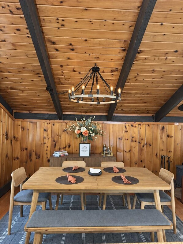 Una habitación con paneles de madera, una gran mesa de comedor, sillas, un banco, una araña y un jarrón con flores.