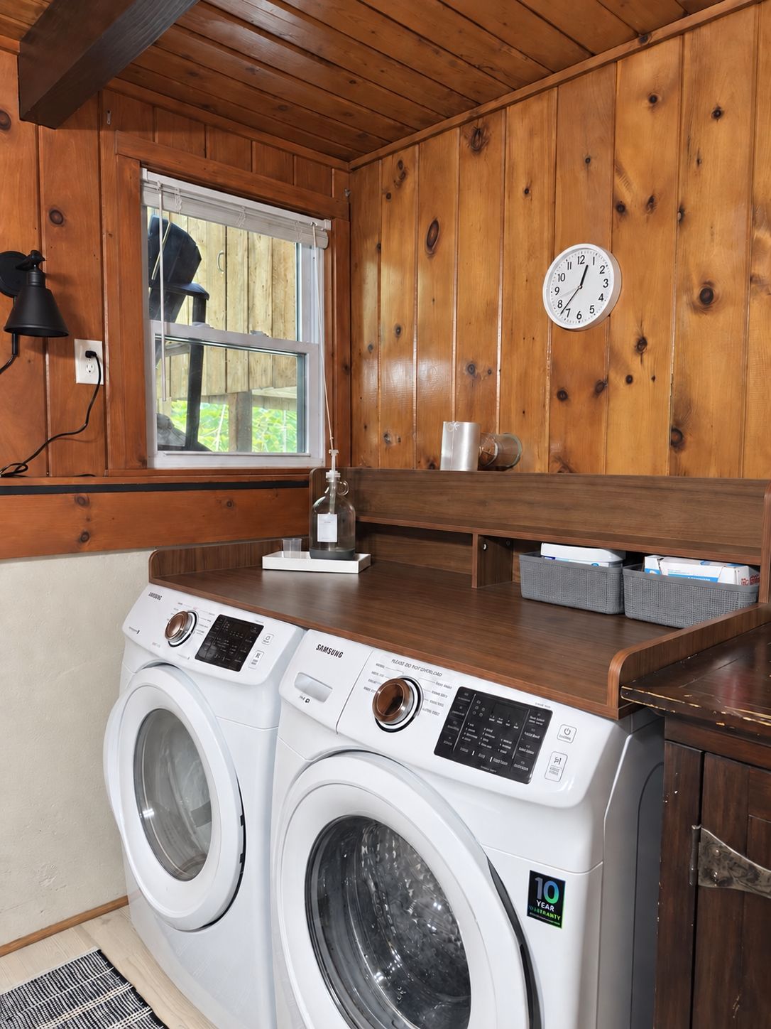 A laundry area with two washing machines, a clock, and a window with a view of greenery.