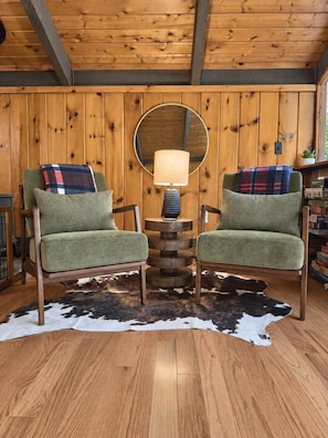 A room with wooden walls, a cowhide rug, two green upholstered chairs, a round table with a lamp, and a mirror on the wall.