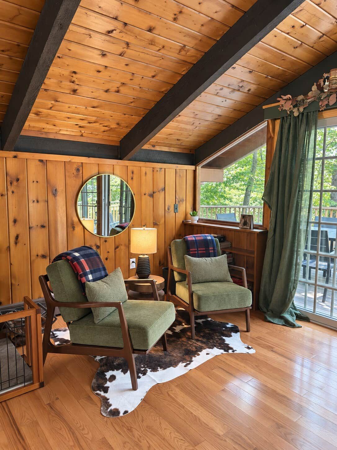 A cozy room with wooden walls and a sloped ceiling, featuring two green armchairs, a round mirror, a small table with a lamp, and a large window with a view of greenery outside.