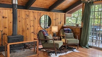 Une pièce chaleureuse aux murs en bois, avec une cheminée, deux fauteuils, un grand miroir rond et une vaste fenêtre agrémentée de rideaux.