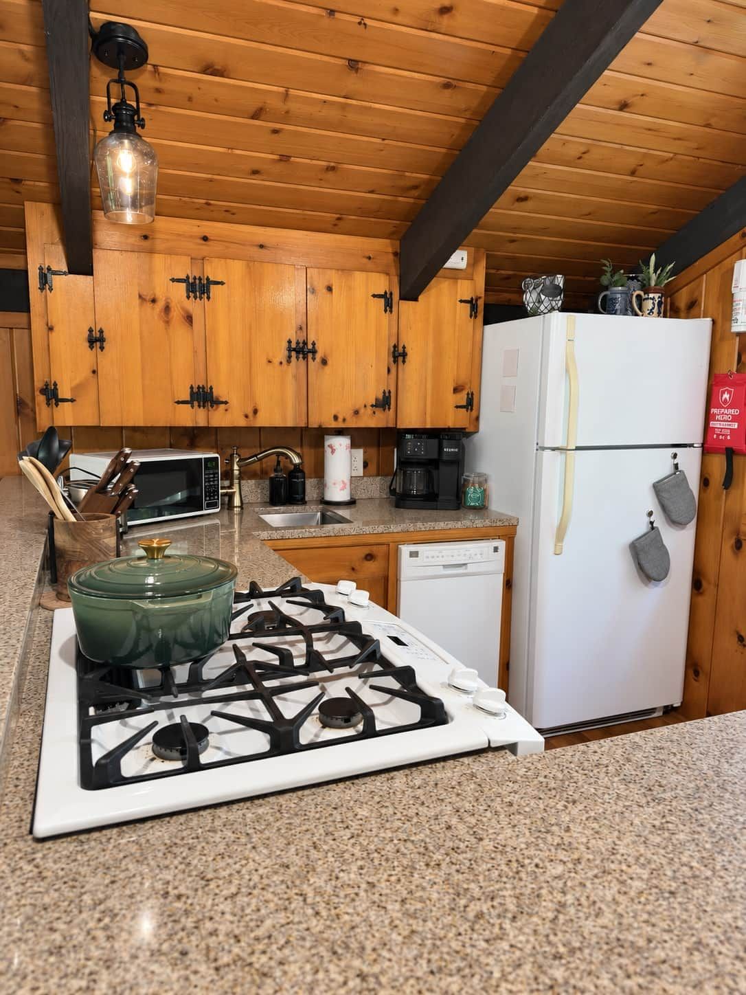 A kitchen with wooden cabinets, a gas stove, a microwave, a refrigerator, and a hanging light fixture.