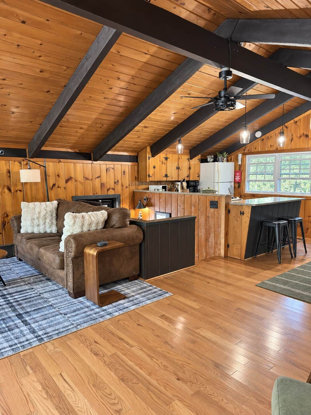 A cozy cabin interior with a wooden ceiling, a sofa, a dining area, and a kitchen.