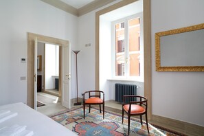 Ein Zimmer mit einem Bett, zwei Stühlen, einem gemusterten Teppich, einem Fenster mit Blick auf Gebäude und einer Heizung.