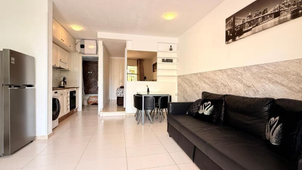 Un salon moderne avec un canapé en cuir noir, une table à manger avec des chaises, un coin cuisine avec un réfrigérateur et une salle de bain avec un miroir.