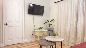 Una habitación con una puerta blanca, un televisor de pantalla plana montado en la pared, una pequeña mesa de madera con una planta en maceta y un sofá rojo.