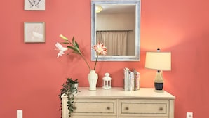 Una habitación con pared rosa, un aparador con cajones, un espejo, un florero con flores, una lámpara y libros.
