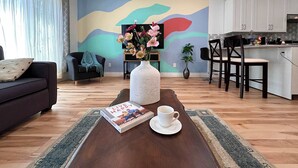Un salon comprenant un canapé, une table basse avec un livre et une tasse, un téléviseur, un mur orné d’œuvres d’art abstrait et un coin cuisine avec des tabourets de bar.