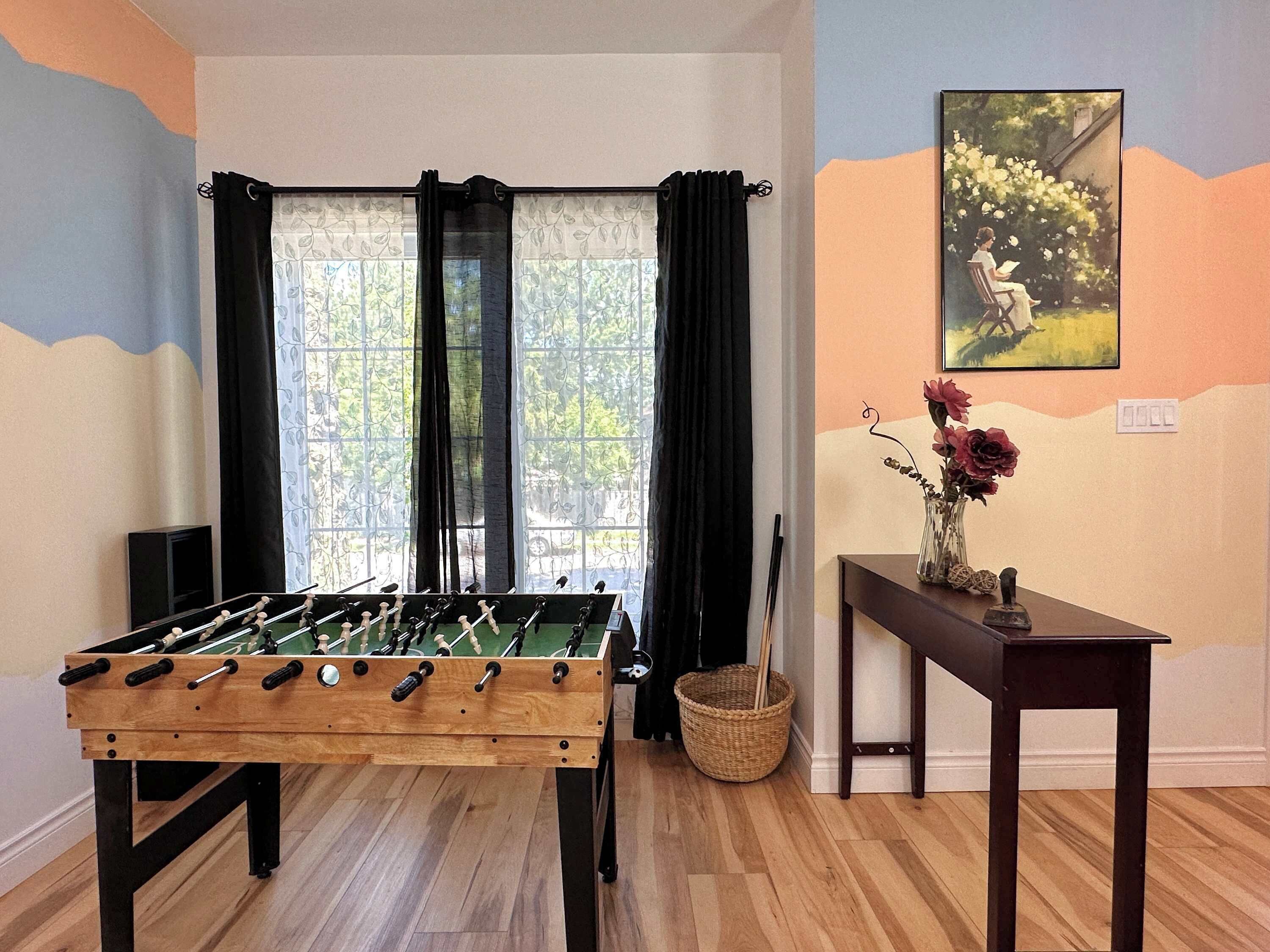 Une pièce avec un baby-foot, une fenêtre avec des rideaux, un tableau accroché au mur et une petite table sur laquelle est posé un vase de fleurs.