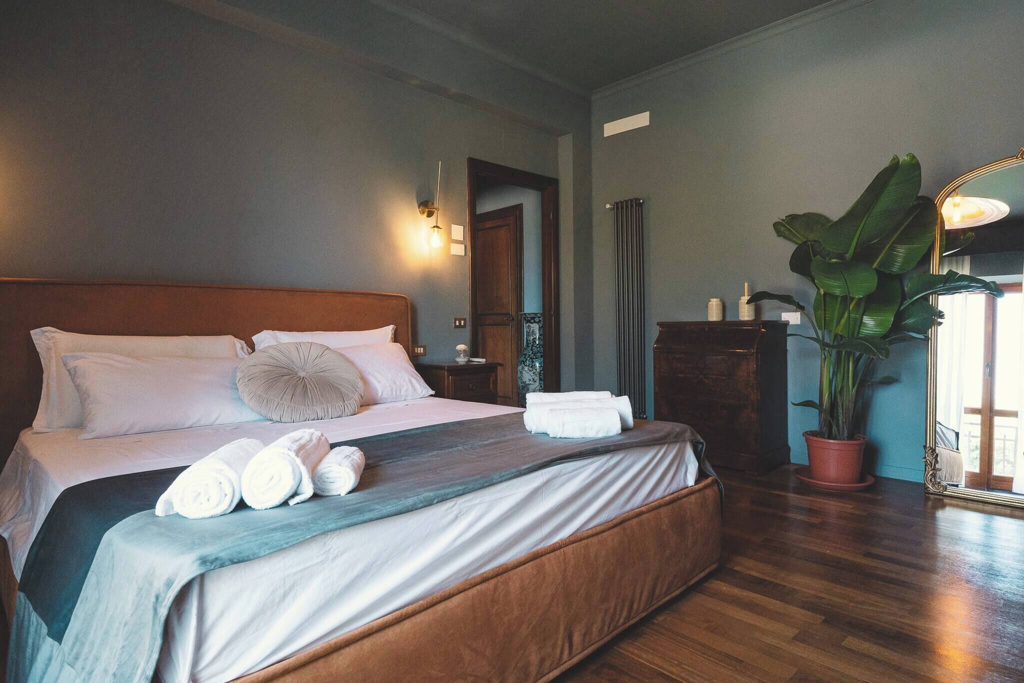 Una cama bien hecha con almohadas y toallas enrolladas, una planta en maceta y un aparador de madera en una habitación con suelo de madera y una puerta que da a otra zona.