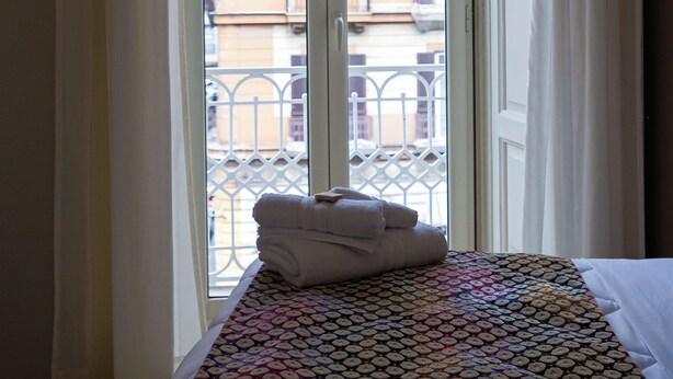 Ein Zimmer mit einem Bett, einem Fenster mit Blick auf Gebäude und einem Balkon mit Geländer.