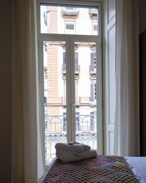 Een kamer met een bed, een raam met uitzicht op gebouwen en een balkon met een leuning.