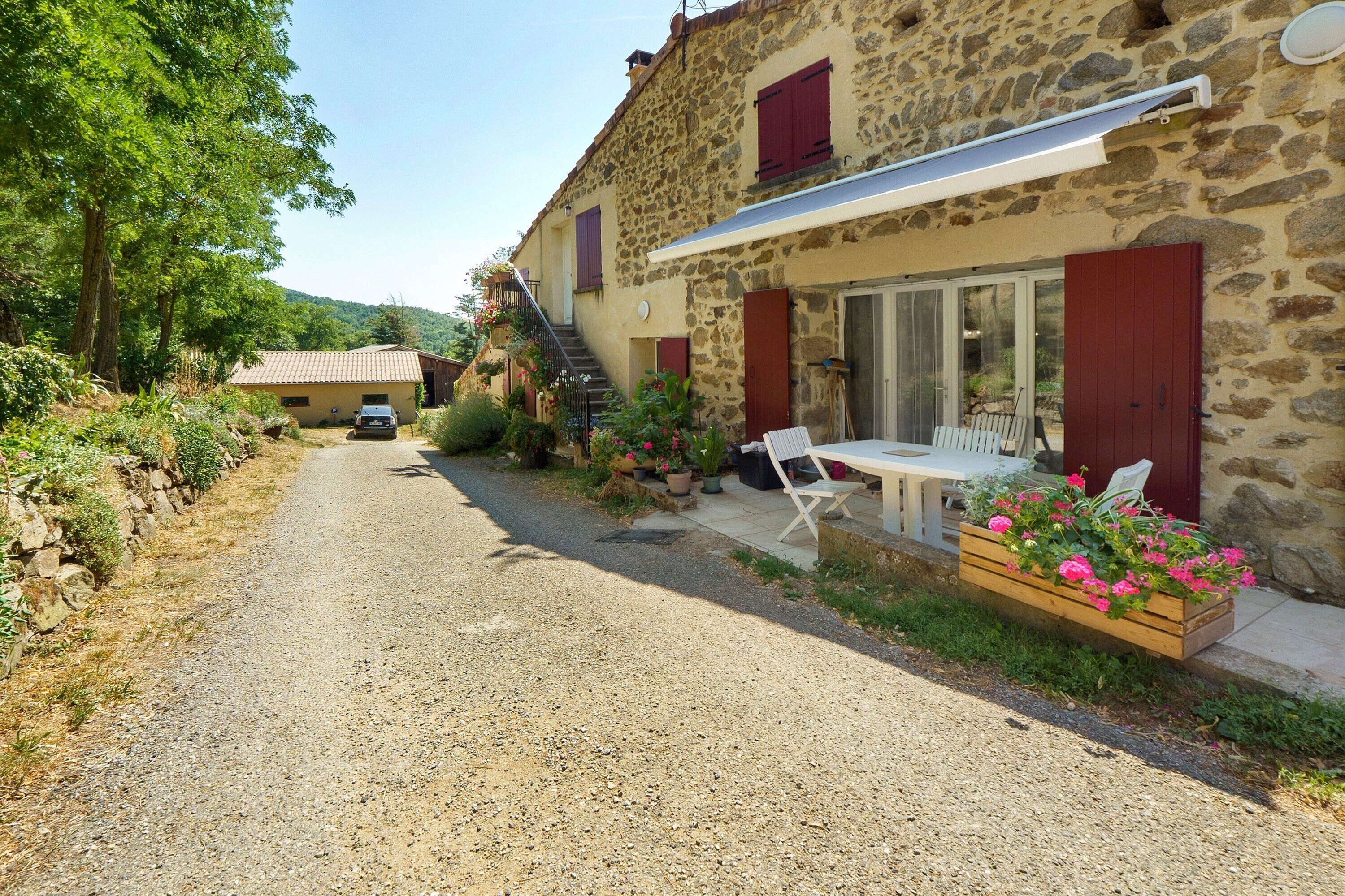 Un bâtiment en pierre avec un chemin de gravier, une voiture garée devant et une table avec des chaises à l’extérieur.