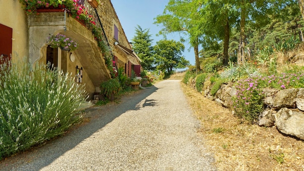 Eine enge Kopfsteinpflasterstraße, gesäumt von Häusern mit Balkonen, die mit Blumen geschmückt sind, führt zu einem üppigen, grünen Hang.