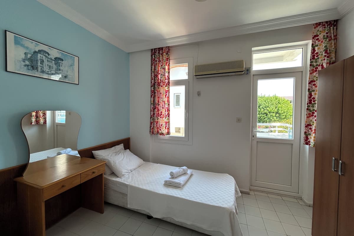 Une chambre avec un lit simple, un bureau en bois, un miroir, une fenêtre avec des rideaux à motifs floraux, un appareil de climatisation et une porte donnant sur un espace verdoyant.