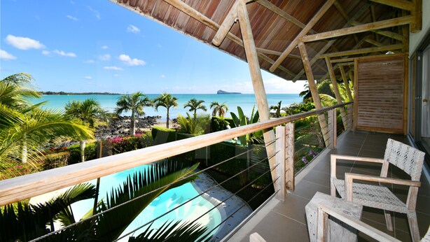 A balcony with a view of a pool and ocean, wooden furniture, and a thatched roof structure. - The Bay Lounge – Beachfront Villa with Pool on a Peninsula ⭐ (Calodyne)