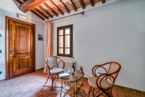 Ein Zimmer mit Holztür, einem Fenster mit Fensterläden, Korbstühlen, einem kleinen Tisch mit Buch und Glas sowie einem Heizkörper.
