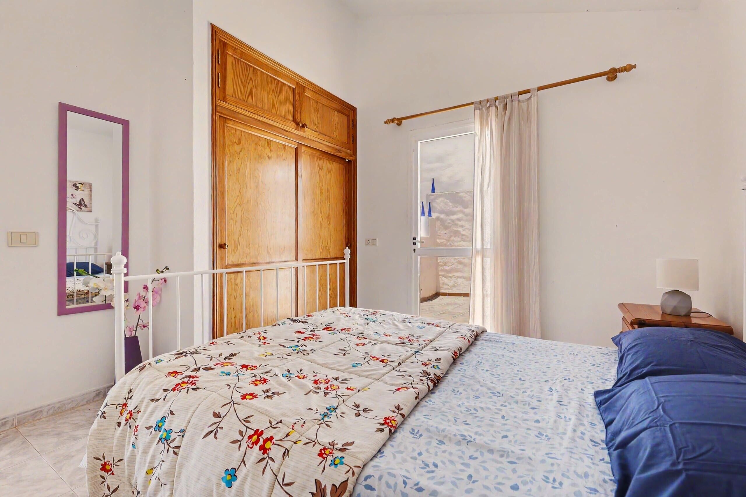 Una camera da letto con un letto, un armadio in legno, uno specchio, un comodino con una lampada e vista all'esterno da una finestra.