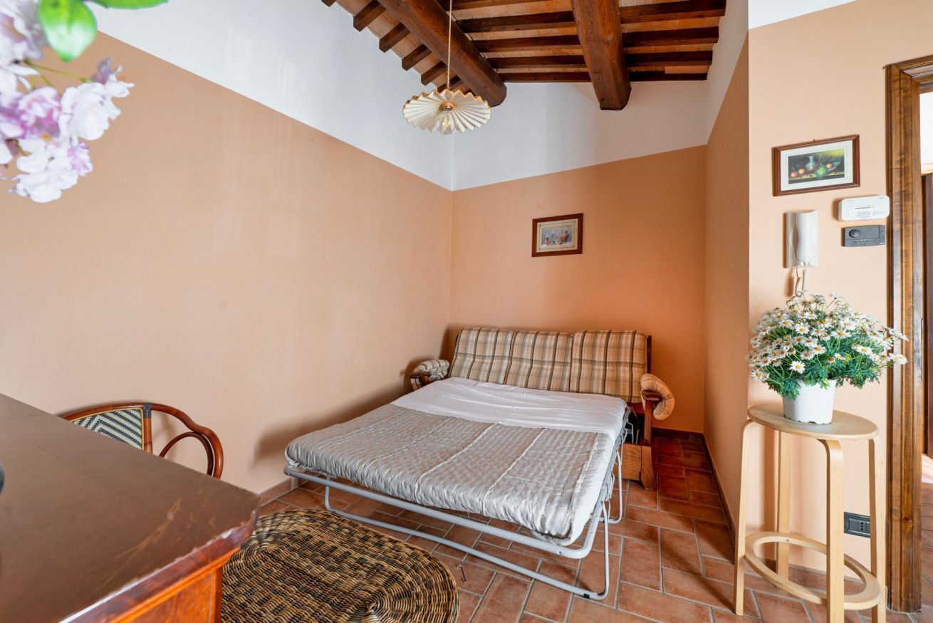 A bedroom with a bed, a wooden table, a wicker chair, a small stool, and a potted plant.