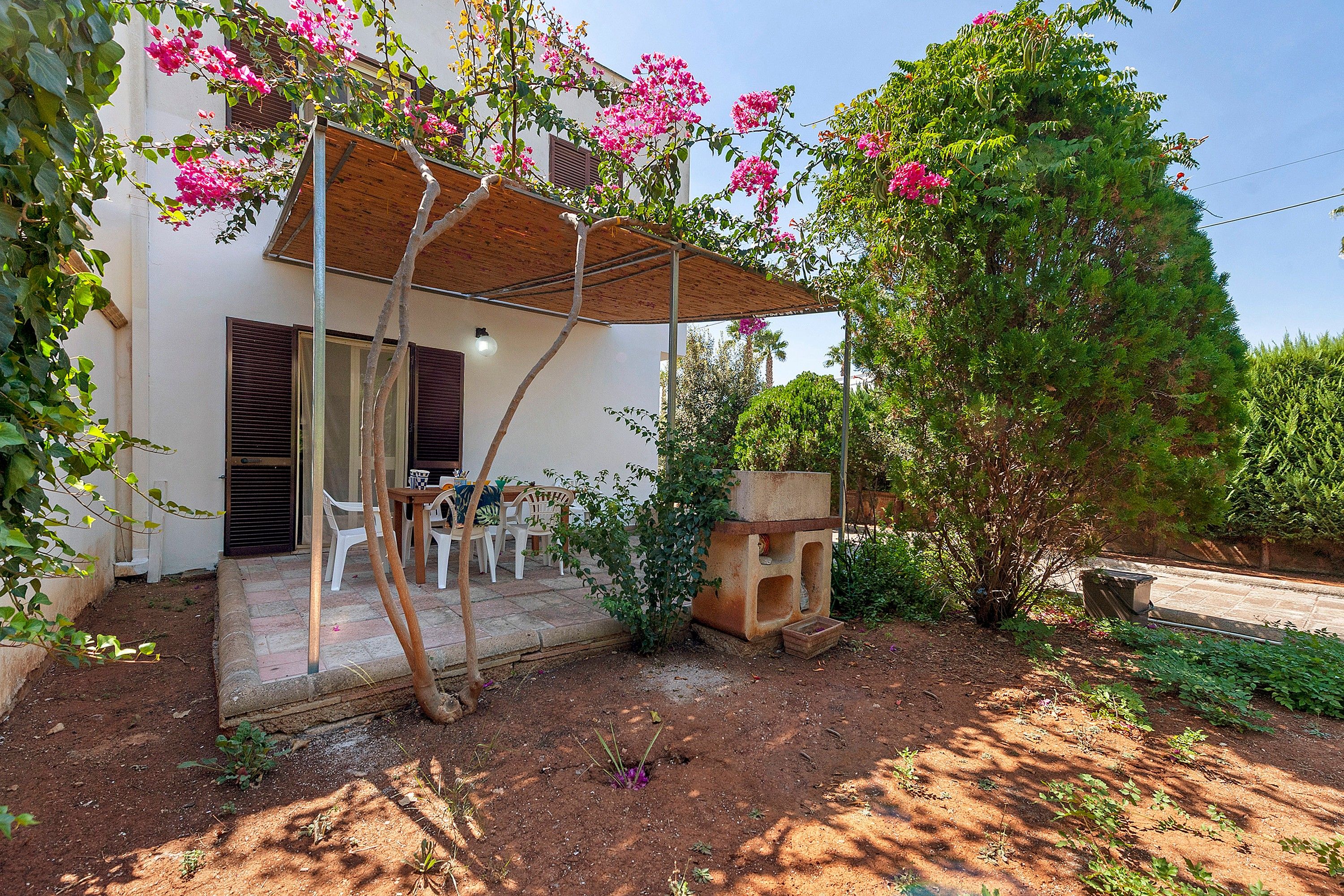 A patio area with a table and chairs, a wooden pergola, and a garden with flowering plants.