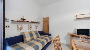 A small, cozy room with a blue sofa, a wooden chair, a dining table, and a television.