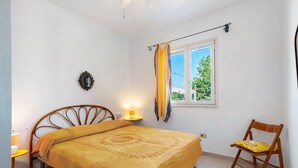 A bedroom with a bed, a chair, a bedside table with a lamp, a window with curtains, and a wall-mounted decoration. - Apartment 'Villetta Sea Sun' with Shared Pool, Private Terrace, and Air Conditioning (Eurovillage)