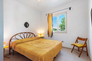 Ein Schlafzimmer mit Bett, Sessel, Nachttisch, Lampe, einem Fenster mit Vorhängen und einer Wanddekoration.