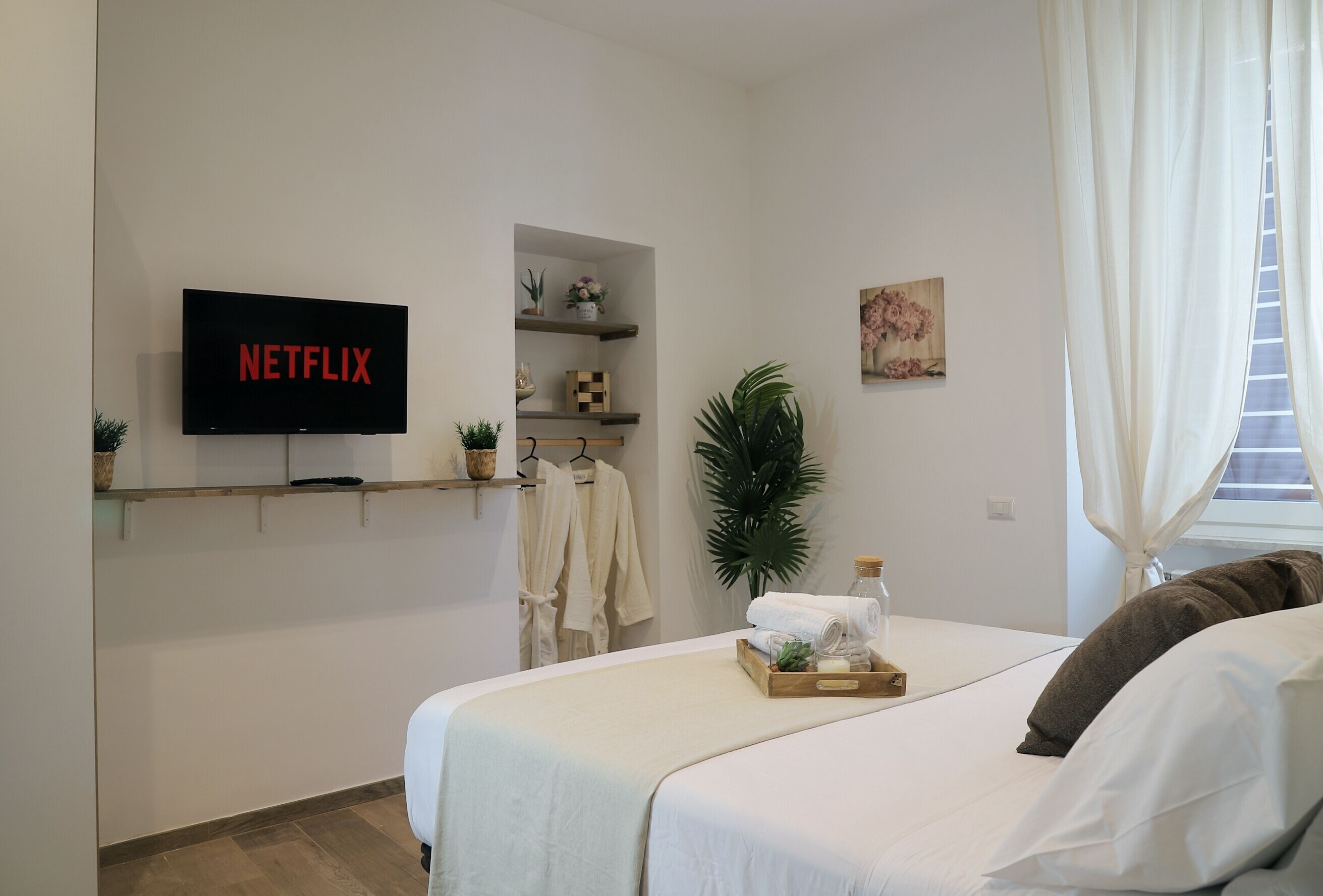 Ein Hotelzimmer mit einem Bett, einem Fernseher, der 'NETFLIX' anzeigt, einer Ablage mit Handtüchern, einer Pflanze und einem Gemälde.