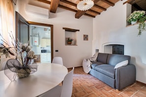 A living room with a sofa, a dining table with chairs, a fireplace, and a kitchen area in the background. - GuestHost - Il Casale dei Gelsi - Narciso (San Martino In Campo)