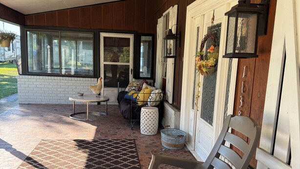 A covered porch with a tiled floor, a rocking chair, a table, and a window with a view of the outdoors.