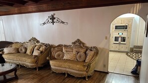 A room with two ornate sofas, a wooden floor, a decorative wall piece, and a glimpse of a kitchen area.