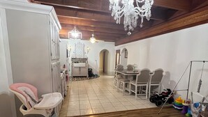 A spacious room with a high ceiling, wooden beams, a chandelier, a dining area with chairs and a table, a baby stroller, and a play area with toys.