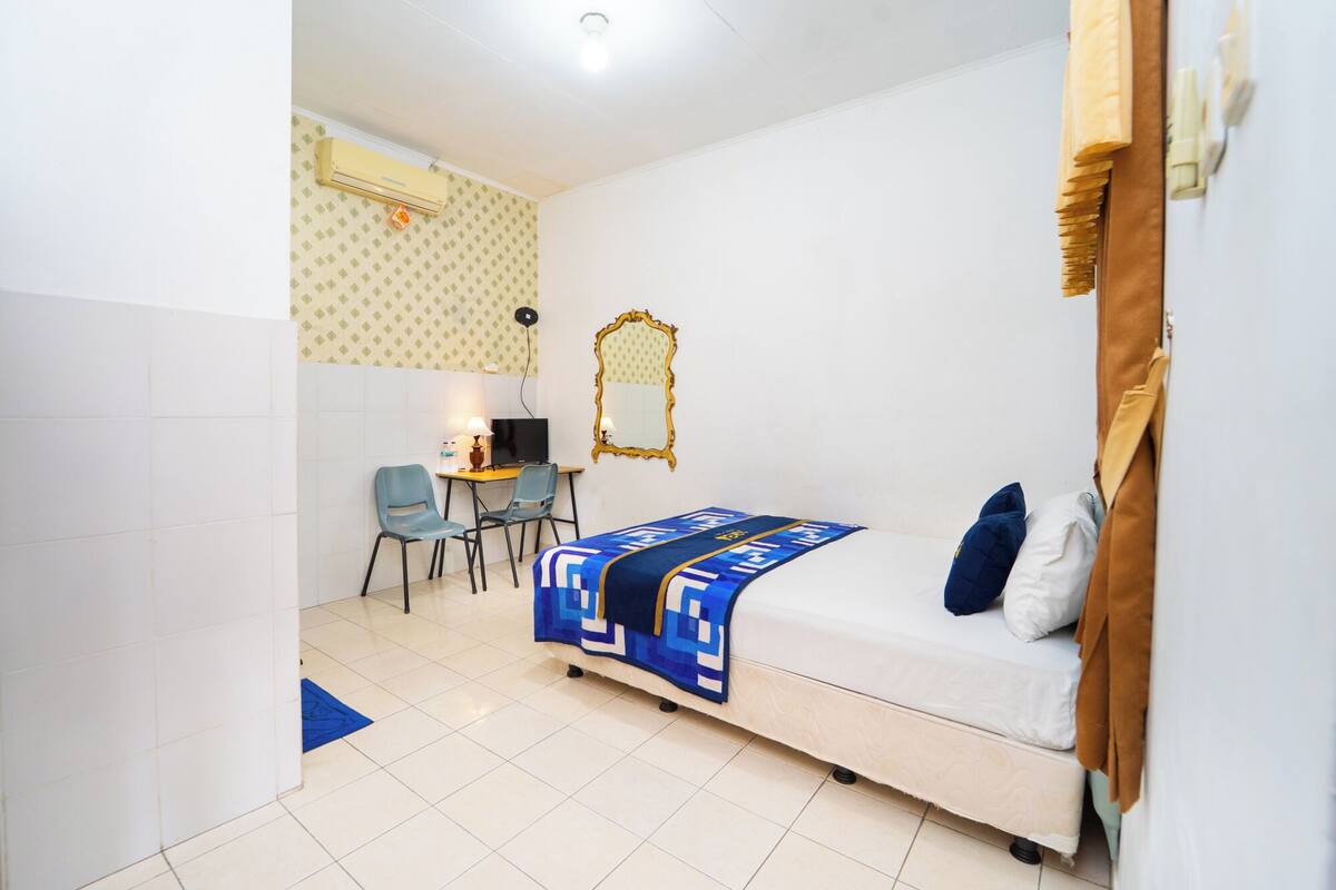 A bedroom with a bed, a desk with a chair, a TV, and an air conditioning unit.
