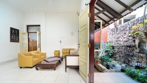 Una habitación con un sofá amarillo, una mesa de centro de madera, una puerta que da a una pared de piedra con un elemento acuático, y un reloj en la pared.