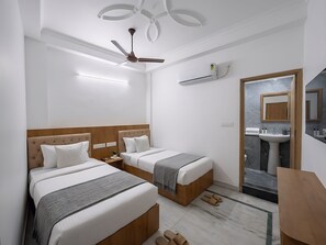 Habitación de hotel con dos camas, ventilador de techo, unidad de aire acondicionado, baño con lavamanos y espejo, y piso de baldosas de mármol.