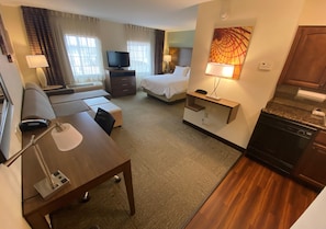 Ett hotellrum med en säng, en soffa, ett skrivbord med en lampa, en tv, en tavla och en liten köksdel.