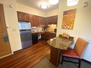 A kitchen with wooden cabinets, a stainless steel refrigerator, a granite countertop, and a red chair. - King Suite with Full Kitchen | Free Breakfast (Great Falls)